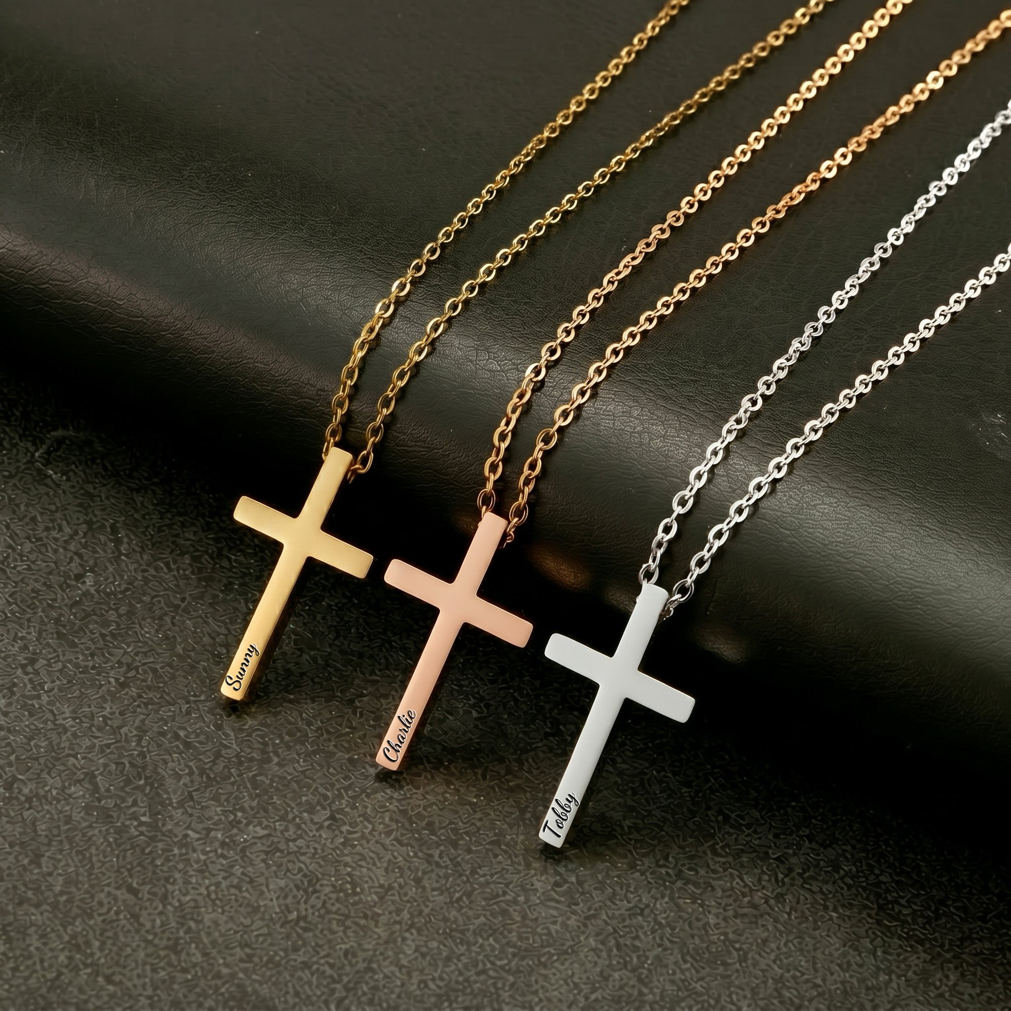 Three personalized cross necklaces in gold, rose gold, and silver, each engraved with a pet name, laid on dark surface