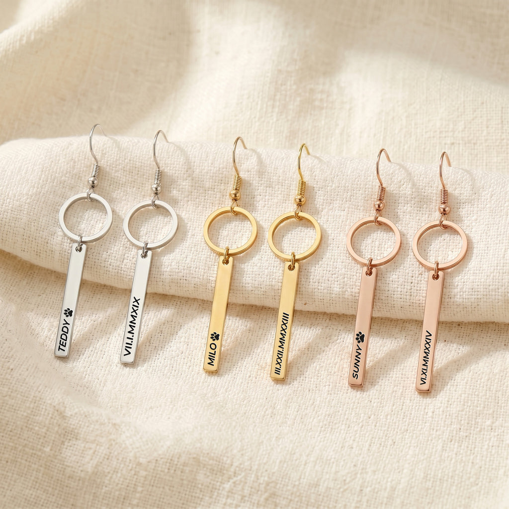 Six pet name bar drop earrings in gold, rose gold, and silver finishes, each engraved with a pet's name, on cream fabric