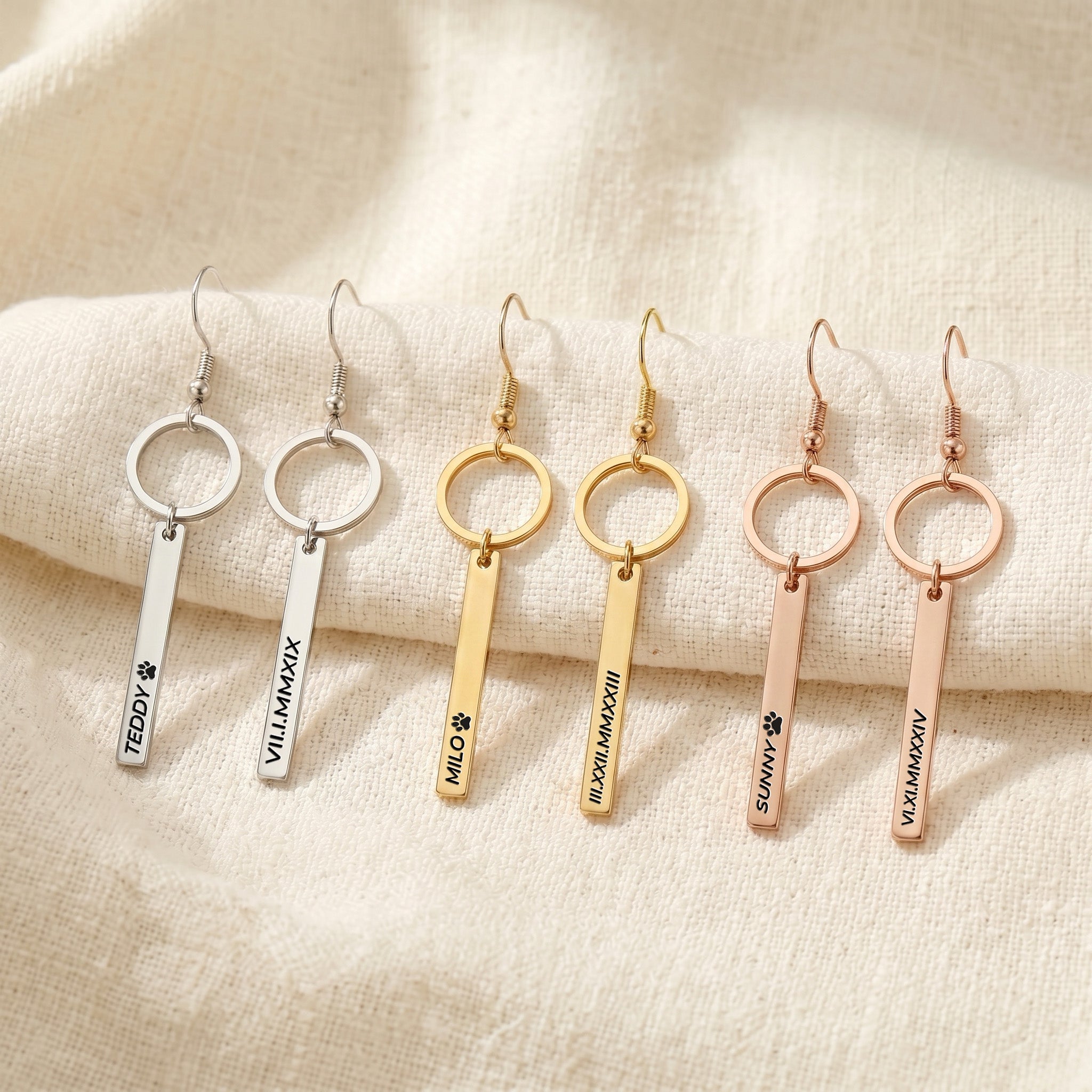Six pet name bar drop earrings in gold, rose gold, and silver finishes, each engraved with a pet's name, on cream fabric