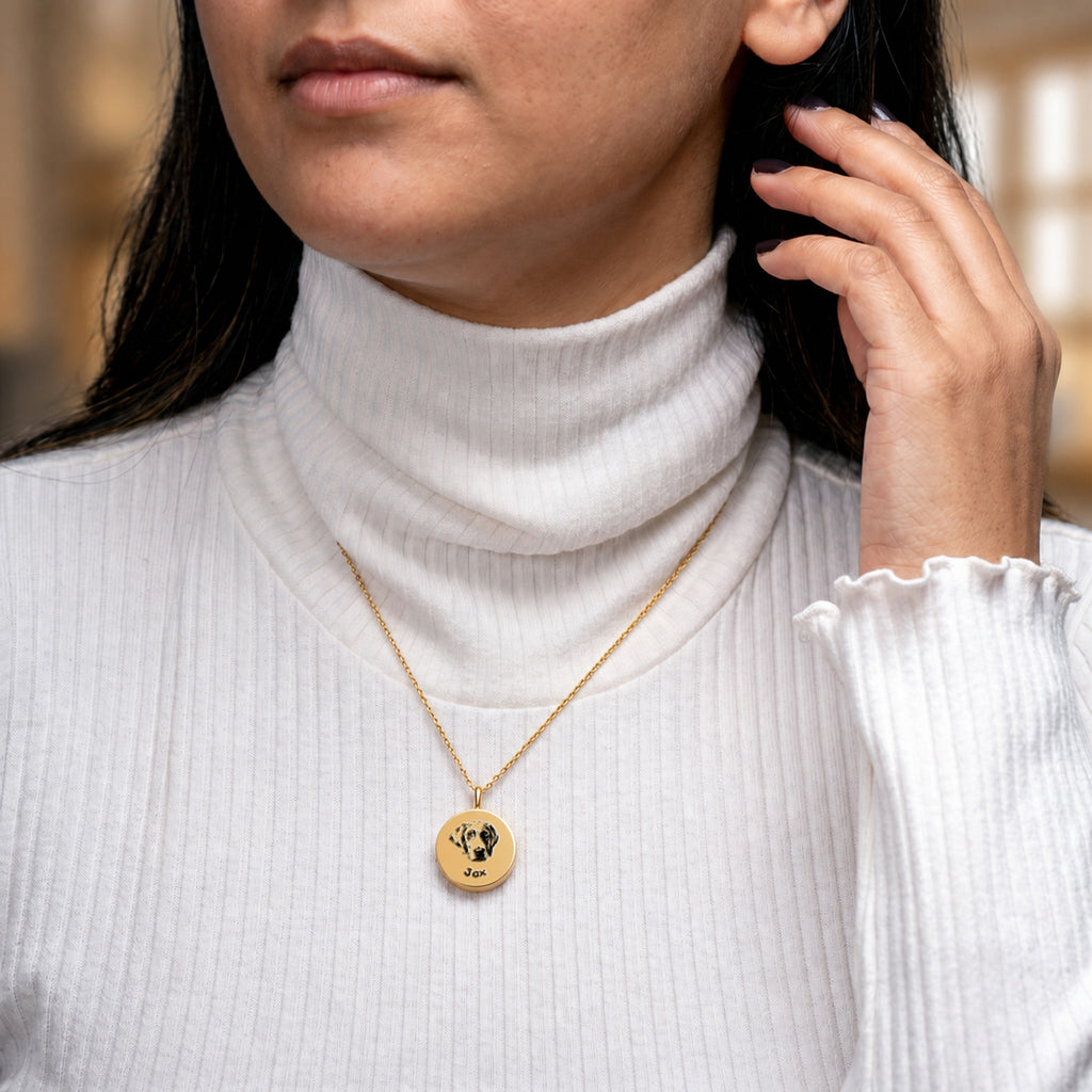 Woman wearing 18K gold pet ashes necklace with engraved dog portrait disc pendant over white turtleneck