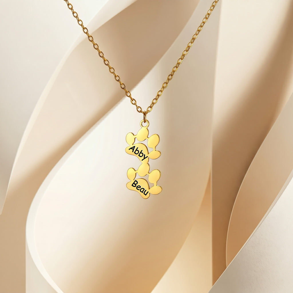 Two gold paw pendants engraved with names on cream fabric flat lay