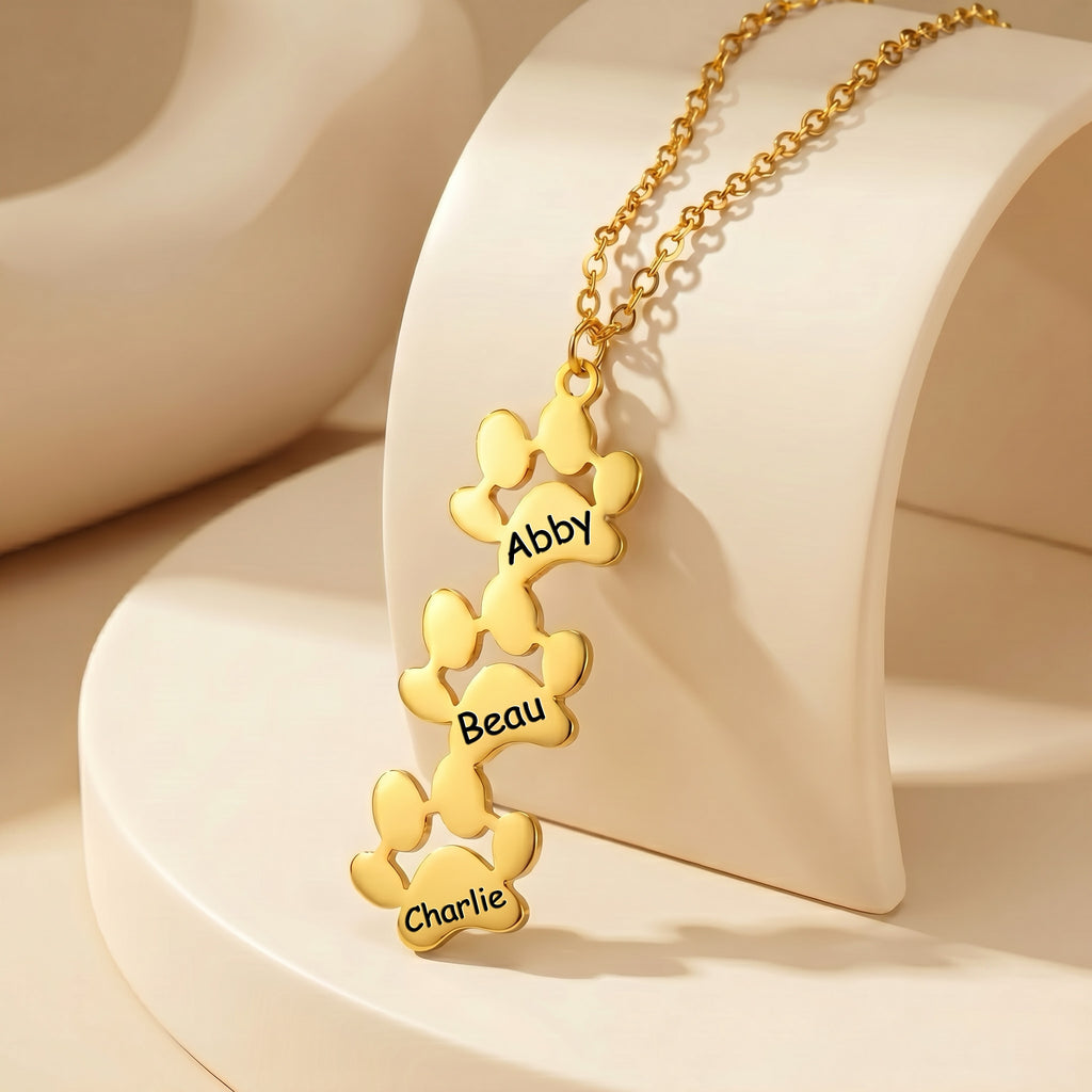 Three gold paw print pendants with engraved names on cream display stand