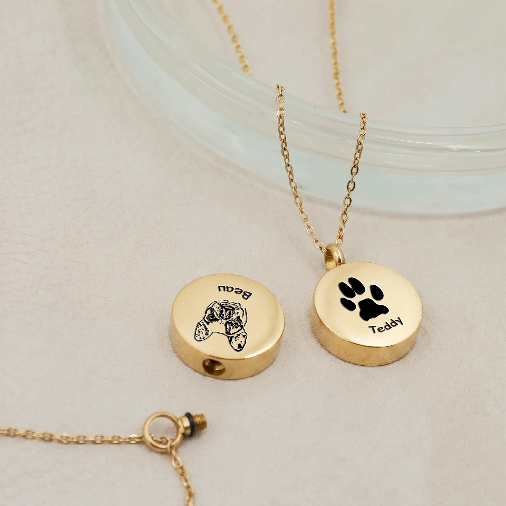 Two pet photo disc urn pendants in 18K gold showing hand-drawn dog portrait and paw print designs with screw cap visible