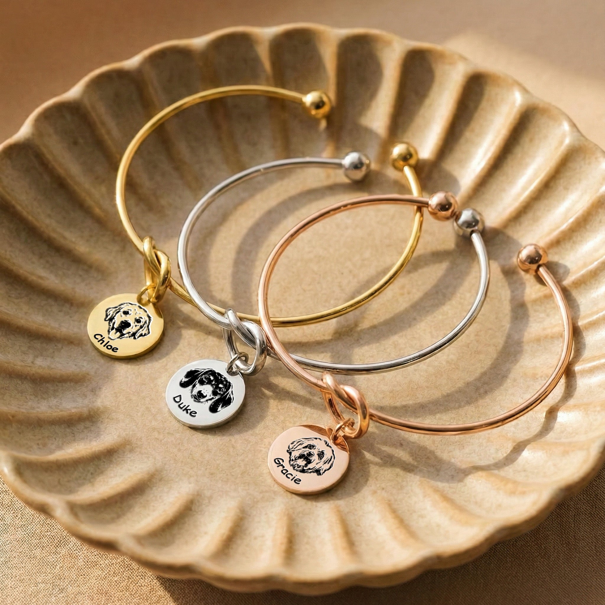 Gold, silver, and rose gold knot cuff bracelets with engraved pet portrait disc charms on a ceramic dish