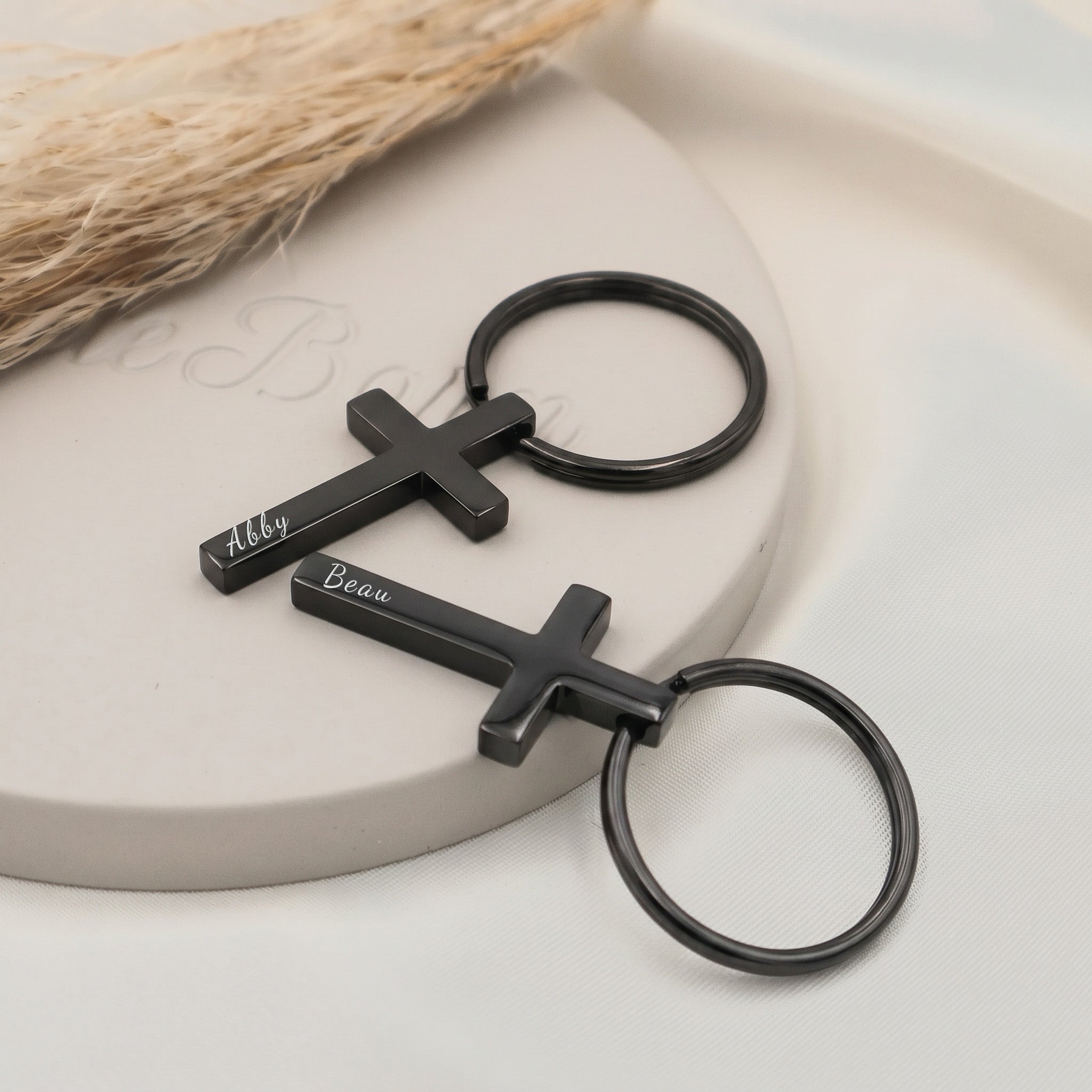 Matte black pet cross keychain shown as a pair staged on a ceramic tray — pet name engraved in script on each cross pendant front (first PupTrace keychain in black plating)