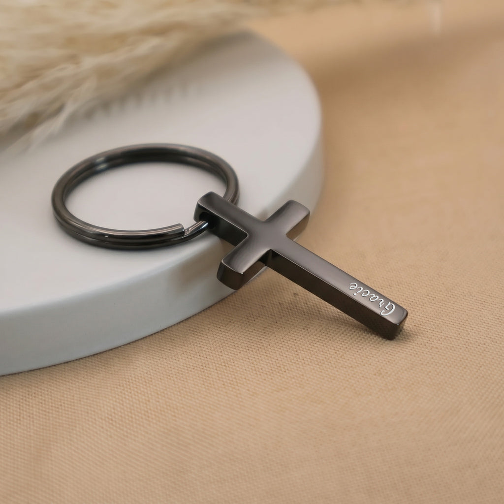 Matte black pet cross keychain angled on a ceramic tray with beige fabric — full pendant and 25mm key ring visible in everyday-carry scene