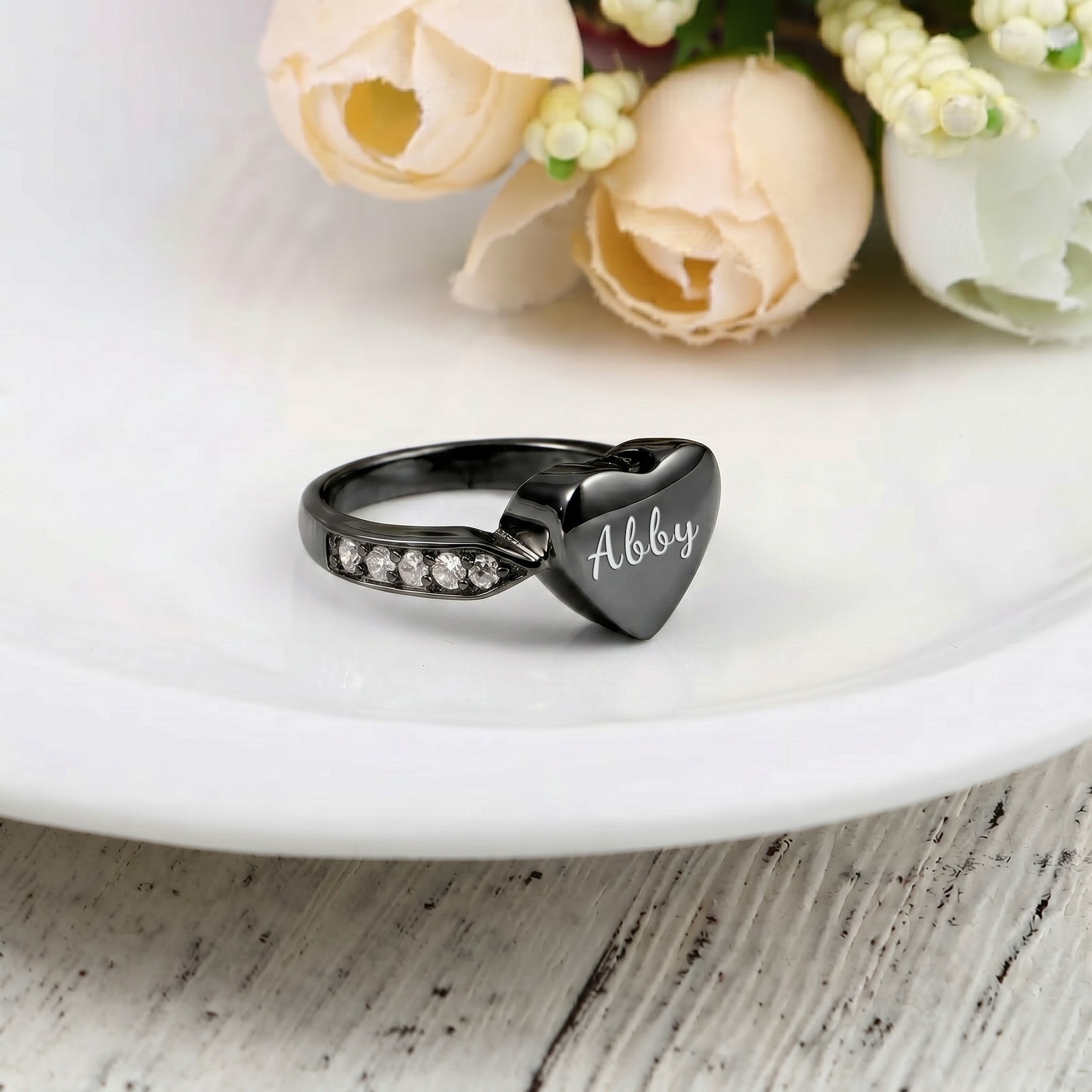 Black heart urn ring with pet name engraved in script, on a white ceramic plate with roses