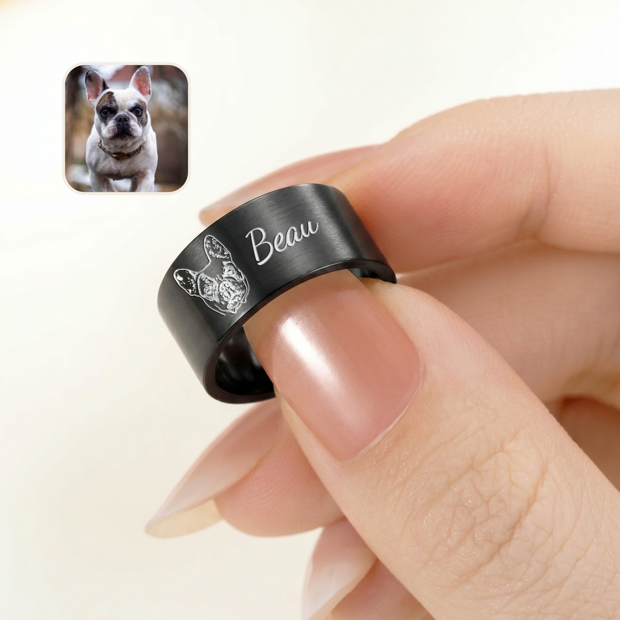 Matte black wide band pet ring pinched between fingers, engraved portrait and script name, with a bulldog reference photo