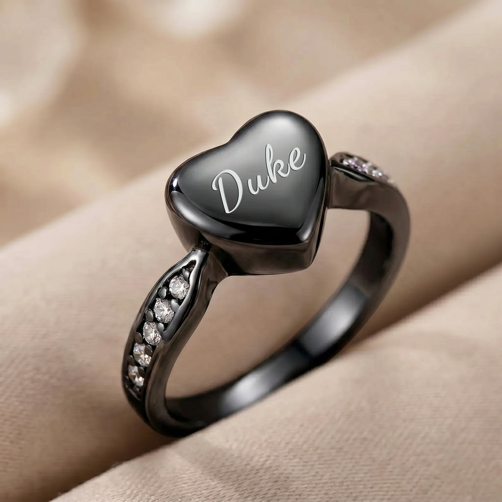 Close-up of black heart urn ring showing white script name and CZ stones against dark plating