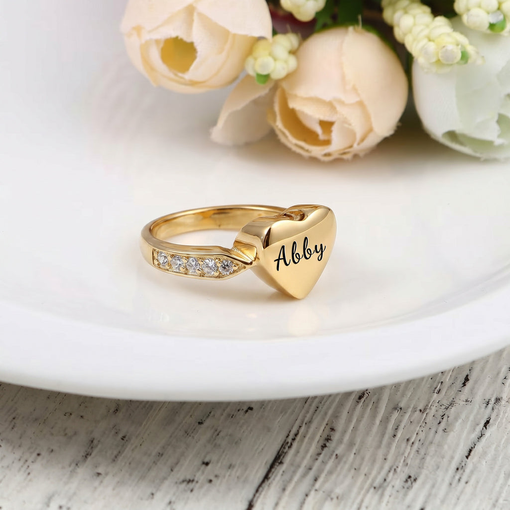 Gold heart urn ring with pet name engraved in script, on a white ceramic plate with roses
