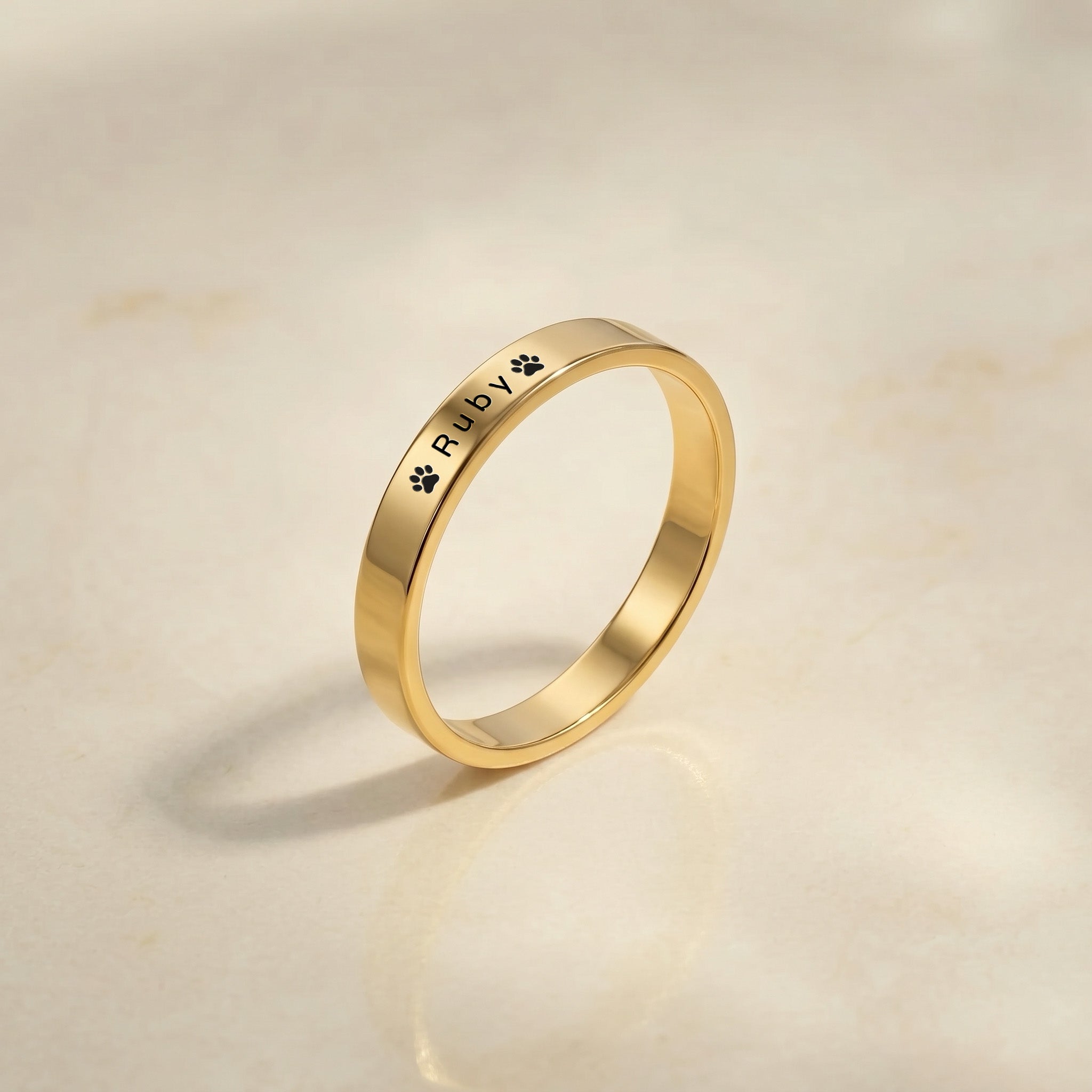 Gold dog name ring with a pet’s name and a paw laser-engraved on a slim band, angled on a cream stone tray