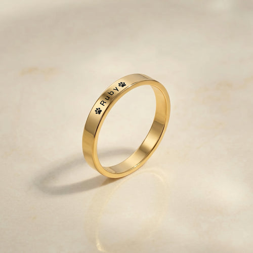 Gold dog name ring with a pet’s name and a paw laser-engraved on a slim band, angled on a cream stone tray