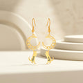 Pair of gold crescent-moon and cat drop earrings with an engraved name, staged upright on a soft beige pedestal
