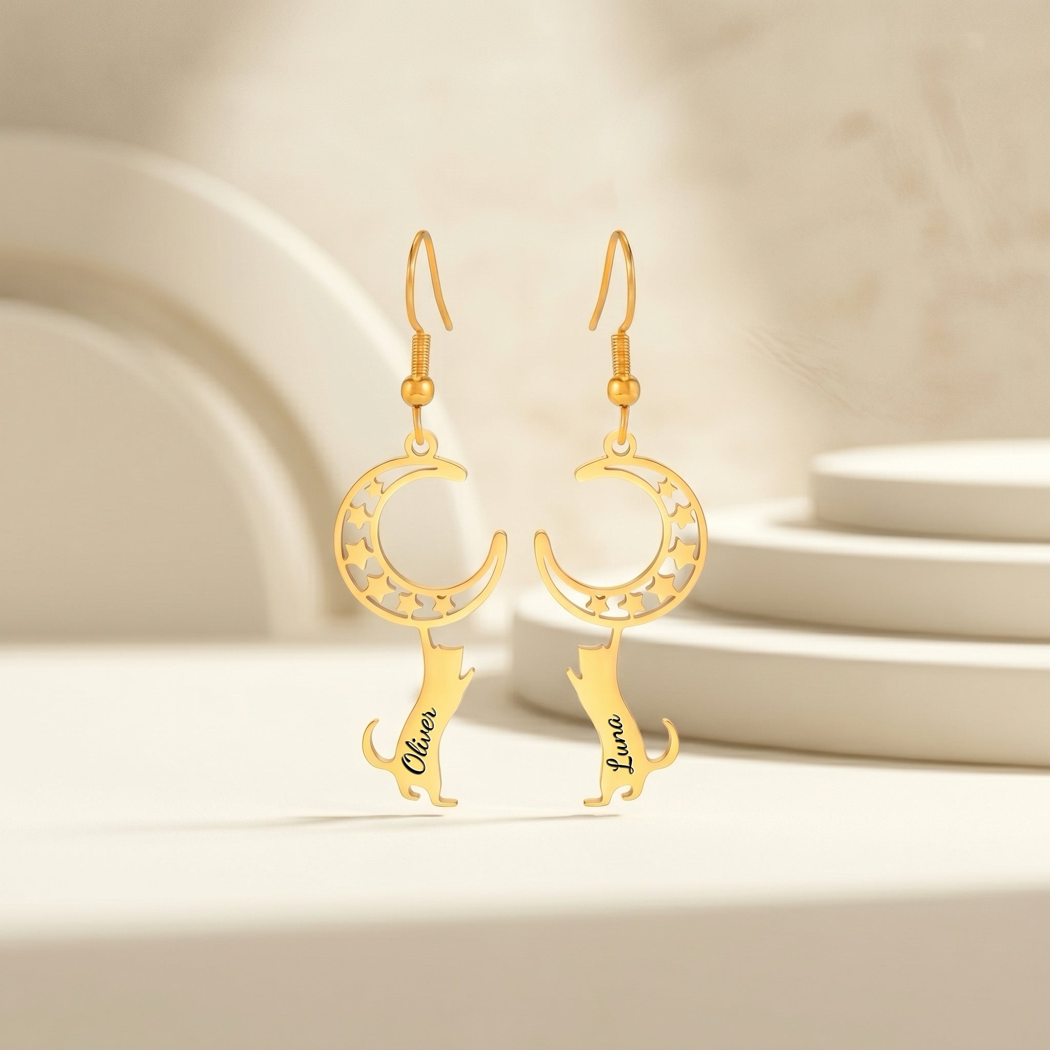 Pair of gold crescent-moon and cat drop earrings with an engraved name, staged upright on a soft beige pedestal