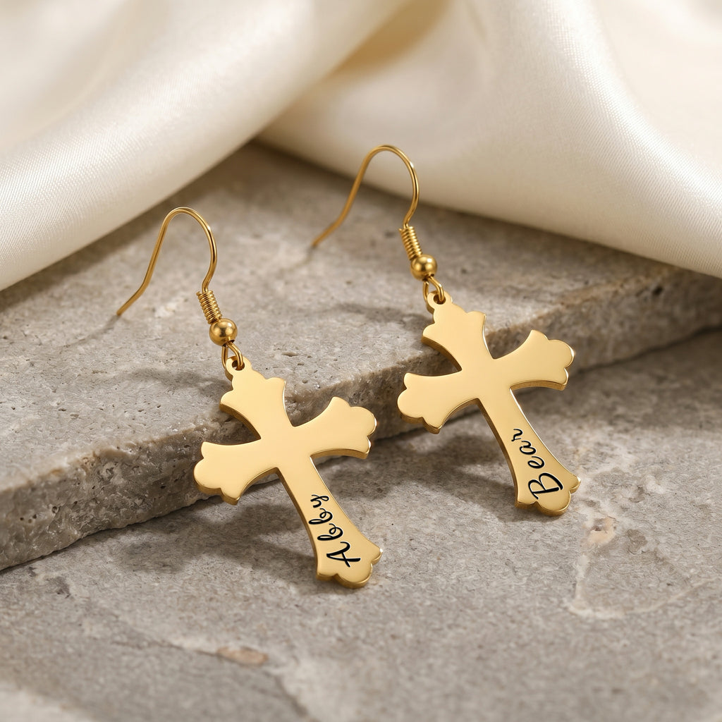 18K gold pet cross earrings shown as a matched pair on a gray stone slab — pet name laser-engraved in script on the lower stem of each cross