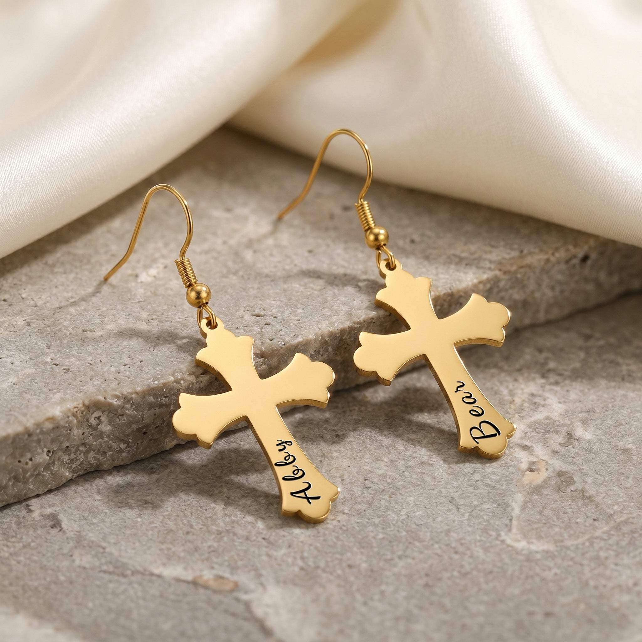 18K gold pet cross earrings shown as a matched pair on a gray stone slab — pet name laser-engraved in script on the lower stem of each cross