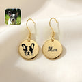Pair of gold drop earrings with hand-drawn dog portrait and name discs, staged on cream fabric beside a photo of the dog