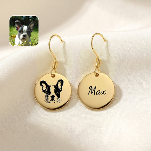 Pair of gold drop earrings with hand-drawn dog portrait and name discs, staged on cream fabric beside a photo of the dog
