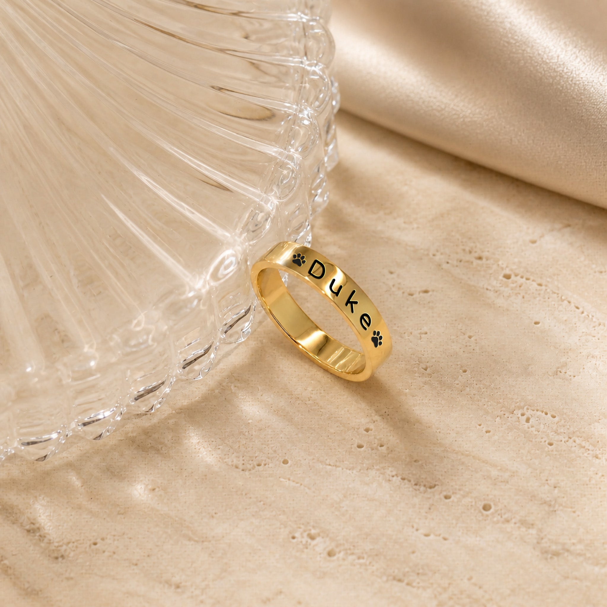 18K gold-plated pet name stacking ring engraved with a name and paw prints, beside a fluted glass dish on travertine