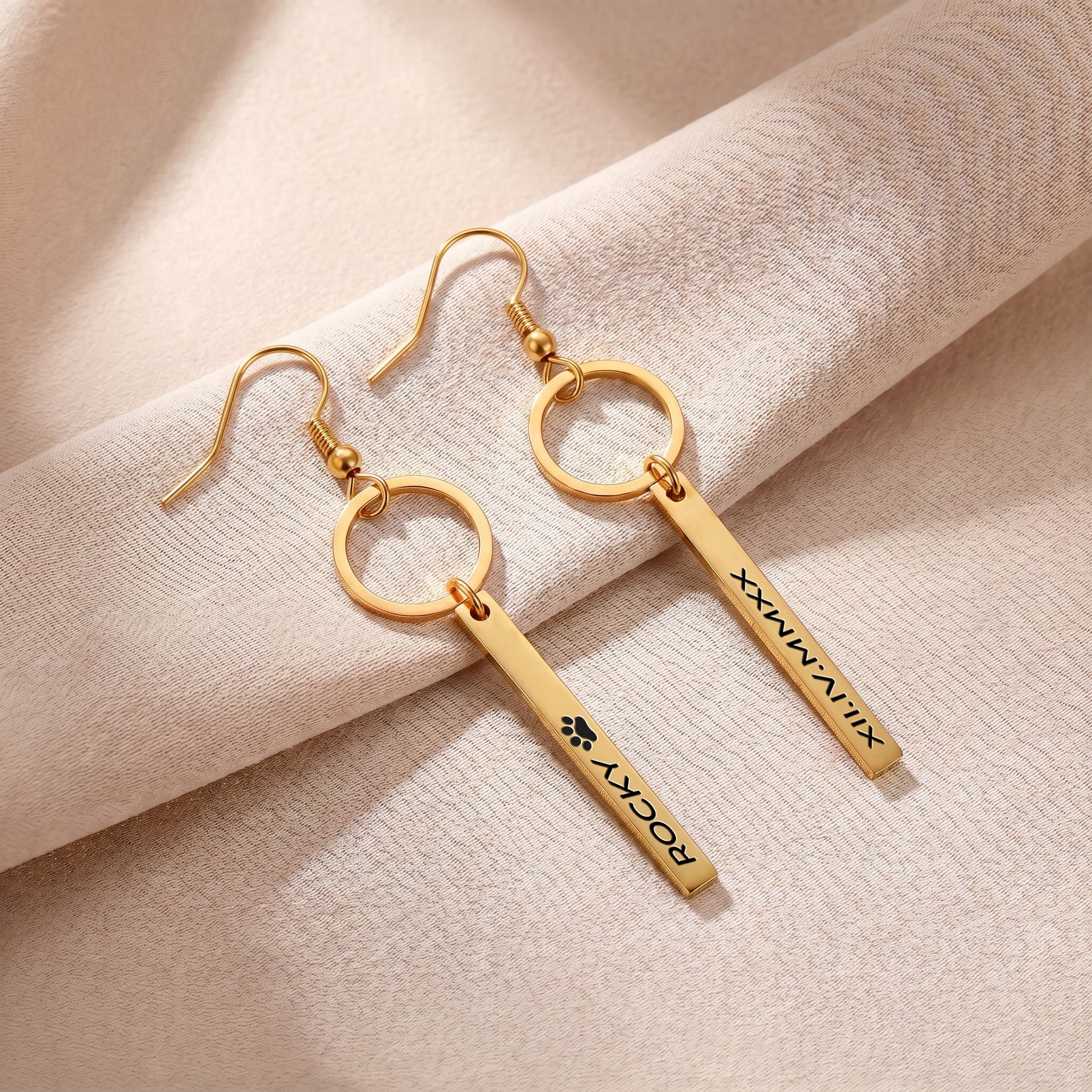 Pair of gold pet name bar drop earrings engraved with a pet's name in block letters, on cream fabric