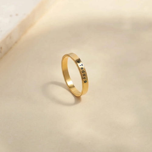 Gold pet name band ring standing on a warm stone surface, name and paw engraving facing forward
