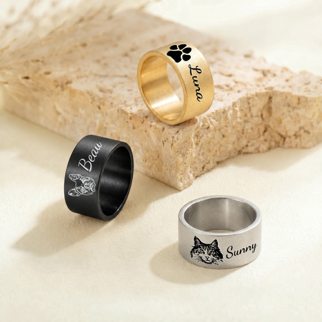 Black, gold and silver wide band pet rings arranged around a beige stone block, showing all three finishes