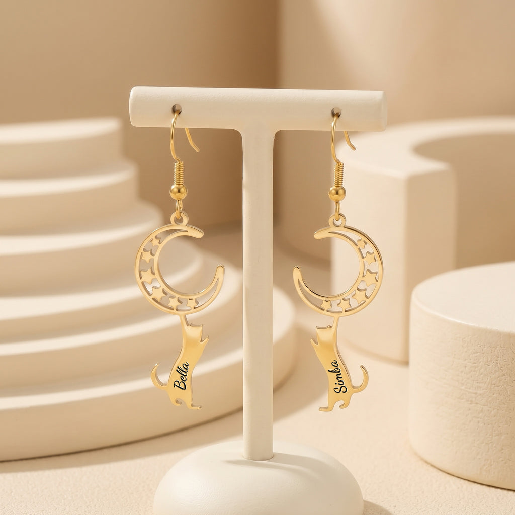 Gold cat-and-moon name earrings hanging from a cream display stand in a neutral ceramic tabletop scene