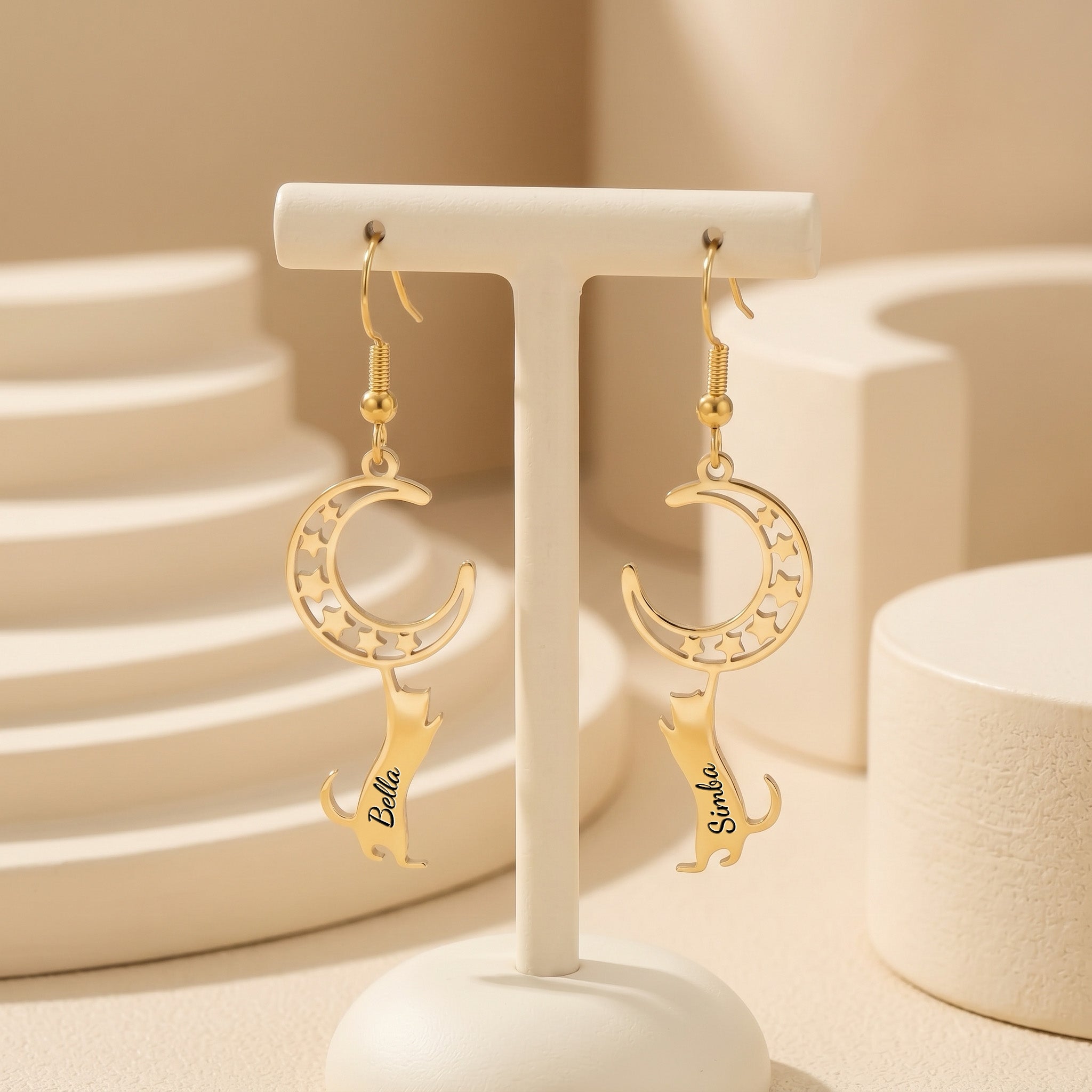 Gold cat-and-moon name earrings hanging from a cream display stand in a neutral ceramic tabletop scene