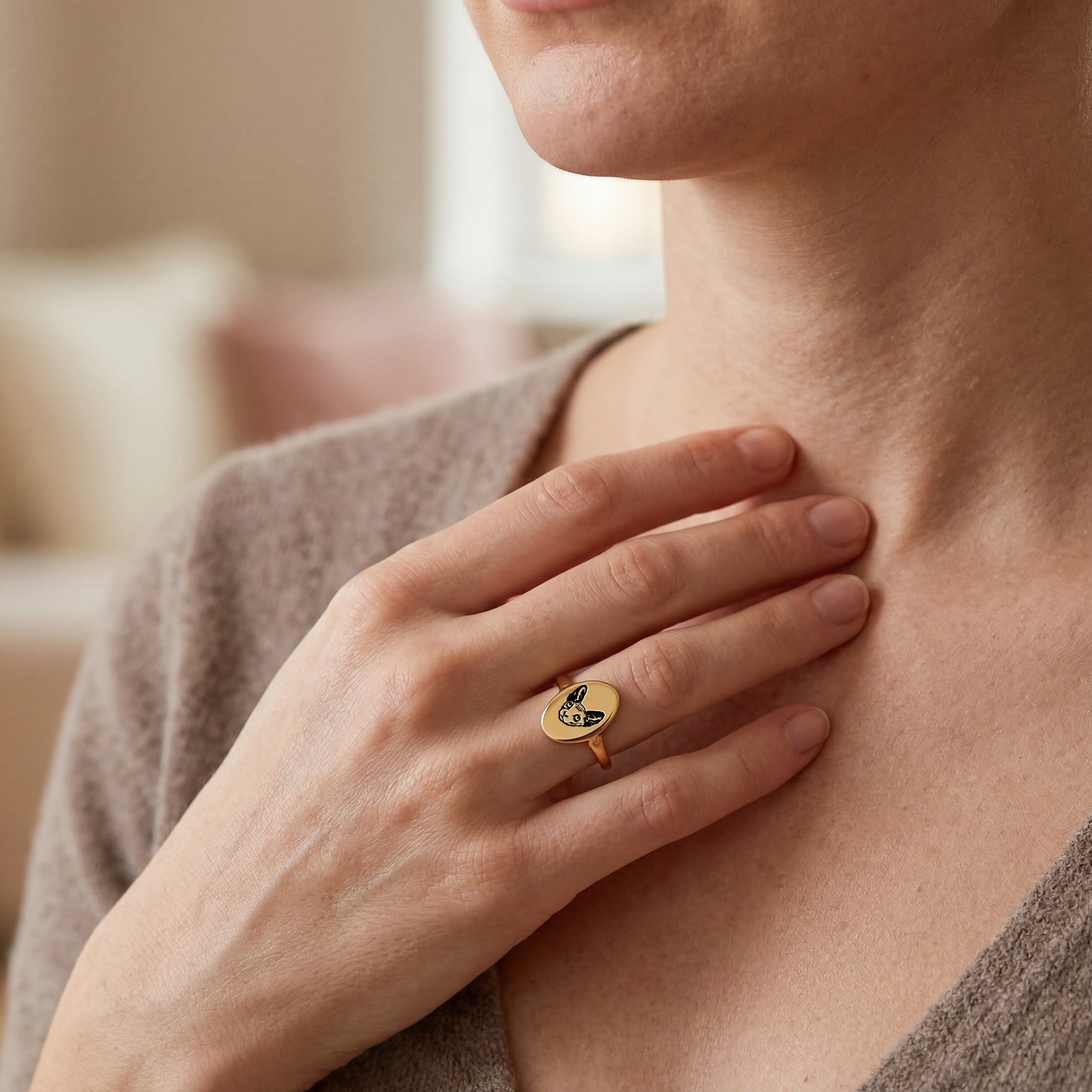 Woman wearing a gold oval signet pet portrait ring, hand resting at her chest in a warm-toned indoor close-up