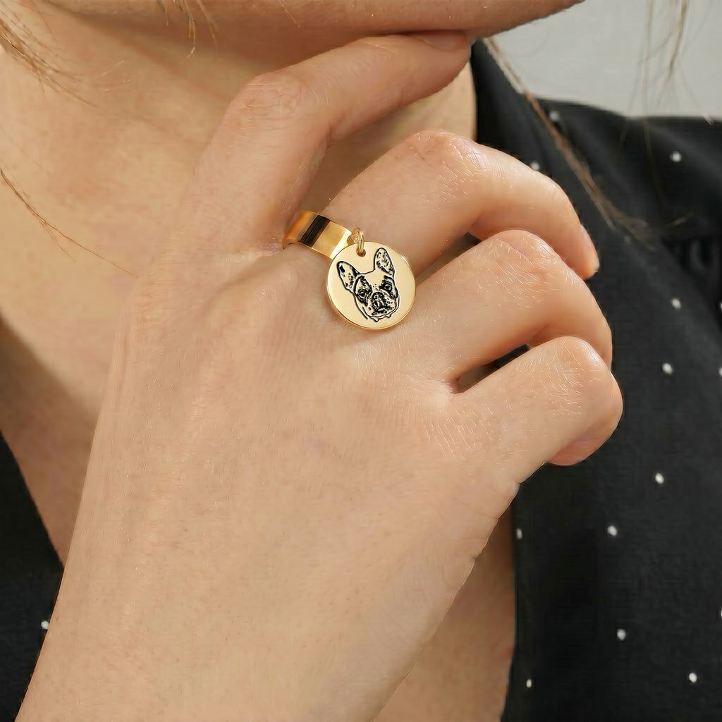 Yellow-gold pet portrait disc ring worn on a finger, held up beside the wearer's face indoors