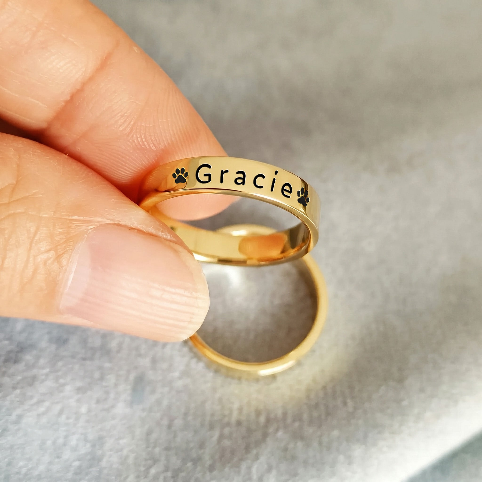 18K gold-plated pet name stacking ring held between fingertips against a soft grey background