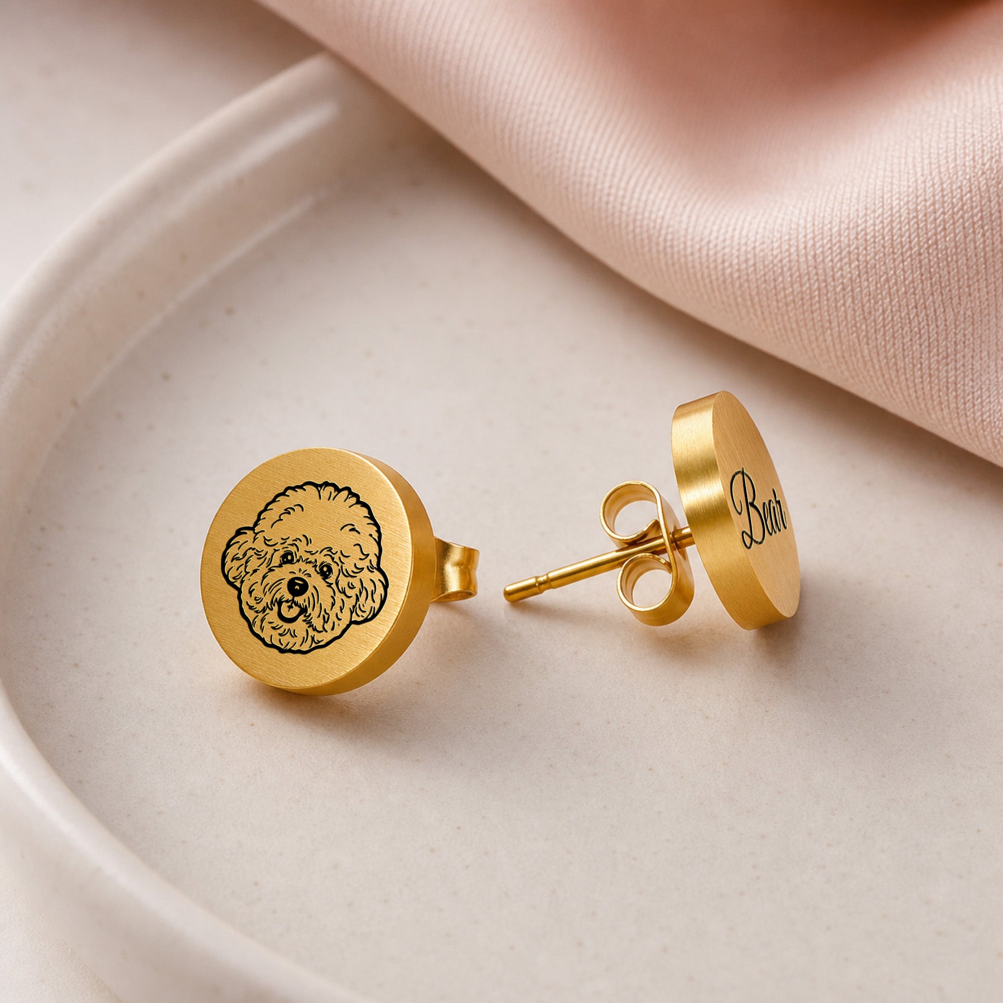 Pair of gold pet portrait stud earrings on a cream dish, hand-drawn portrait stud beside engraved name stud