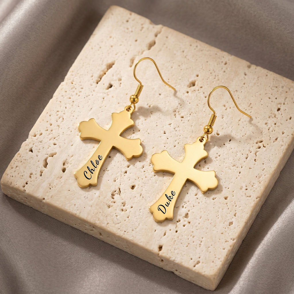 18K gold pet cross earrings, the pair arranged on a cream travertine tile — script pet-name engraving visible on each cross
