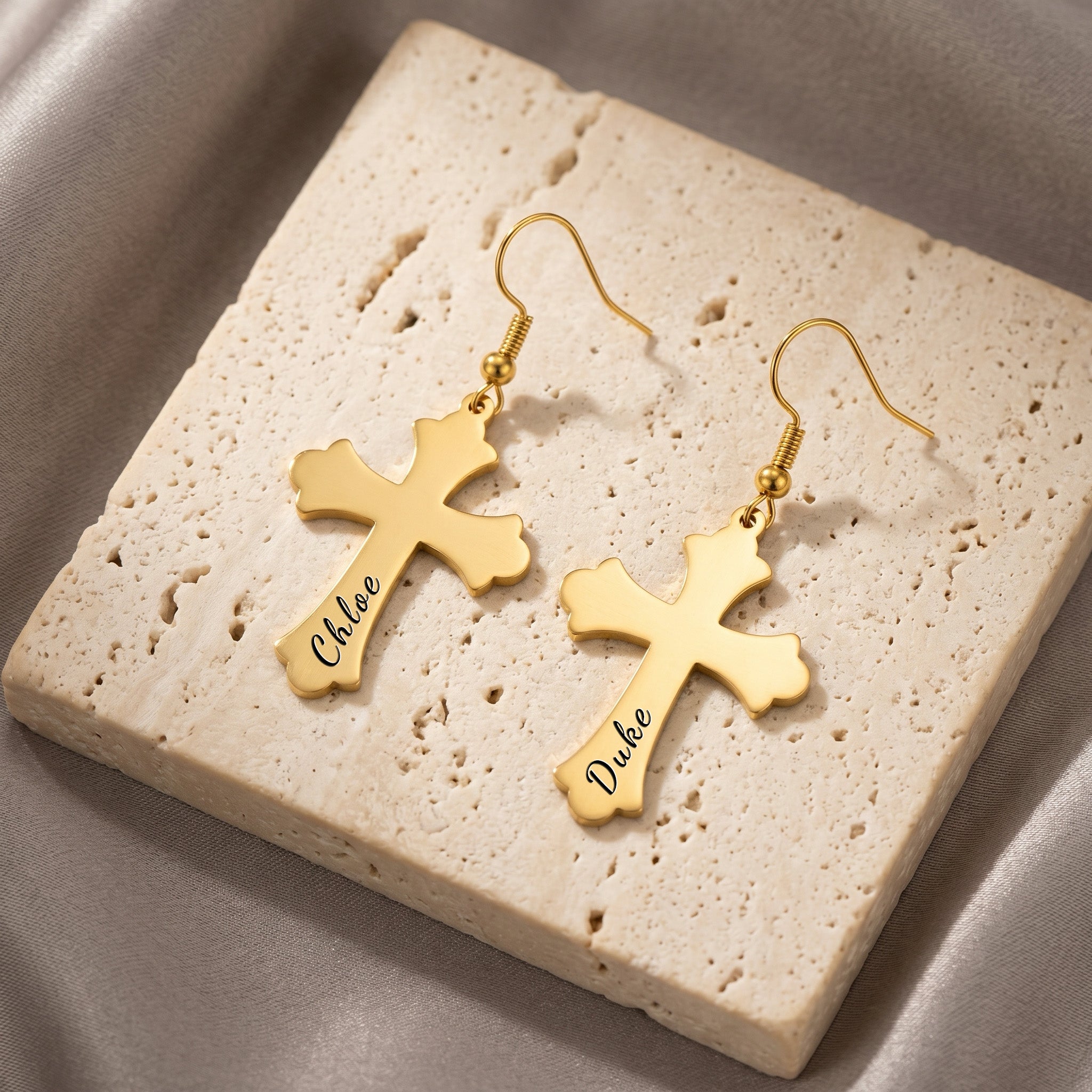18K gold pet cross earrings, the pair arranged on a cream travertine tile — script pet-name engraving visible on each cross