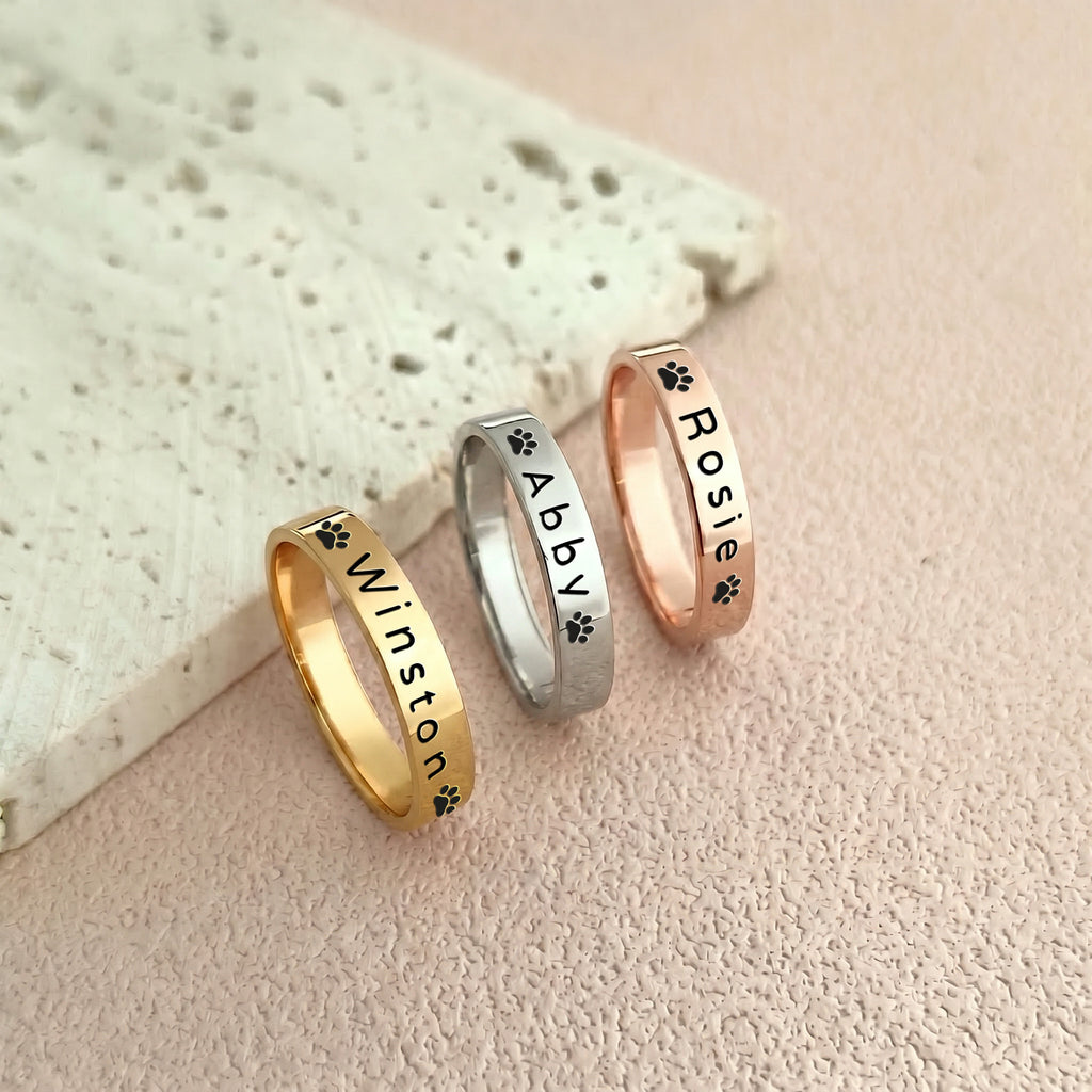 Three pet name stacking rings side by side in 18K gold, rhodium, and 18K rose gold, each engraved with a different name and paw prints