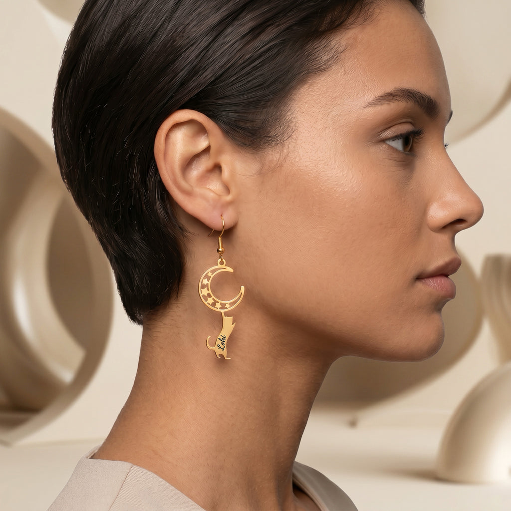 Gold cat-and-moon name earring worn in side profile against a warm neutral sculptural background