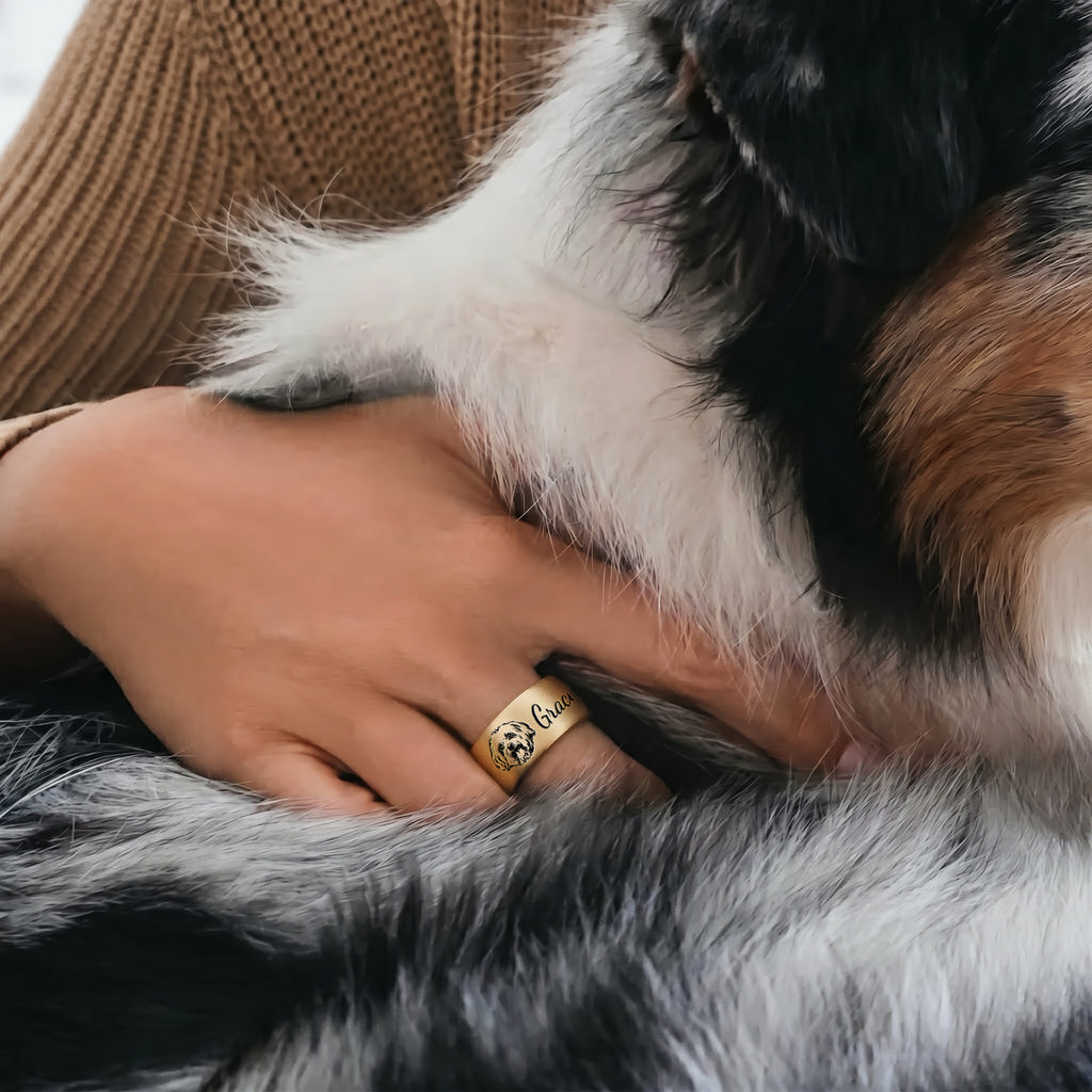 Gold wide band pet ring worn on a finger, engraved portrait and script name, a hand resting near a dog in a home scene