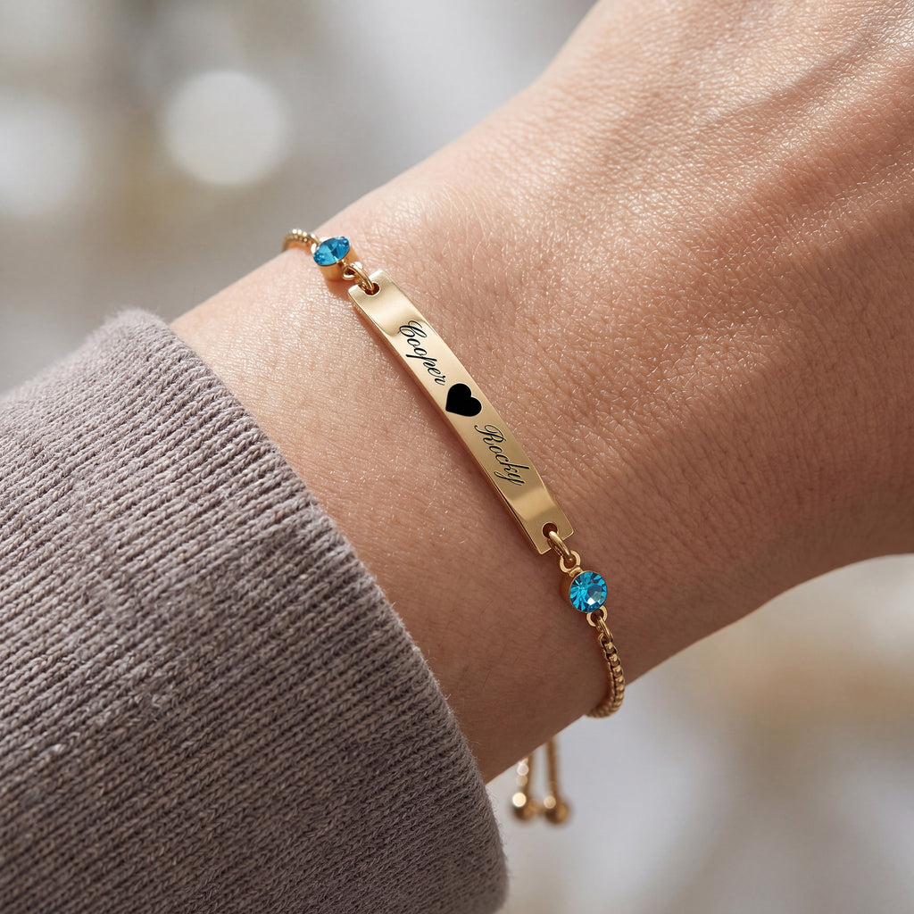 18K gold name bar bracelet on a wrist, engraved text with heart separator and blue birthstones at each end, slider chain