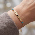 18K gold name bar bracelet on a wrist, engraved text with heart separator and blue birthstones at each end, slider chain