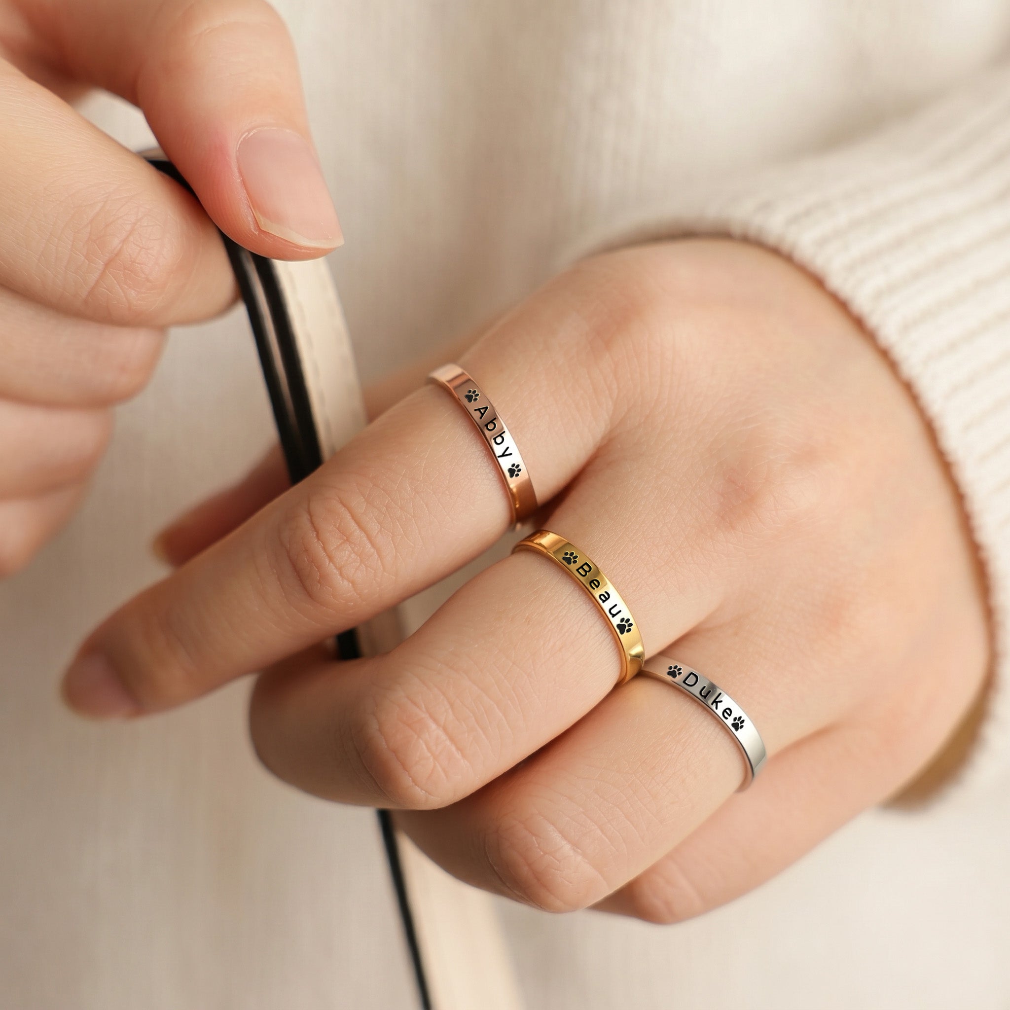 Three slim pet name band rings worn across fingers, the hand holding a cord, in gold, rose gold and silver finishes