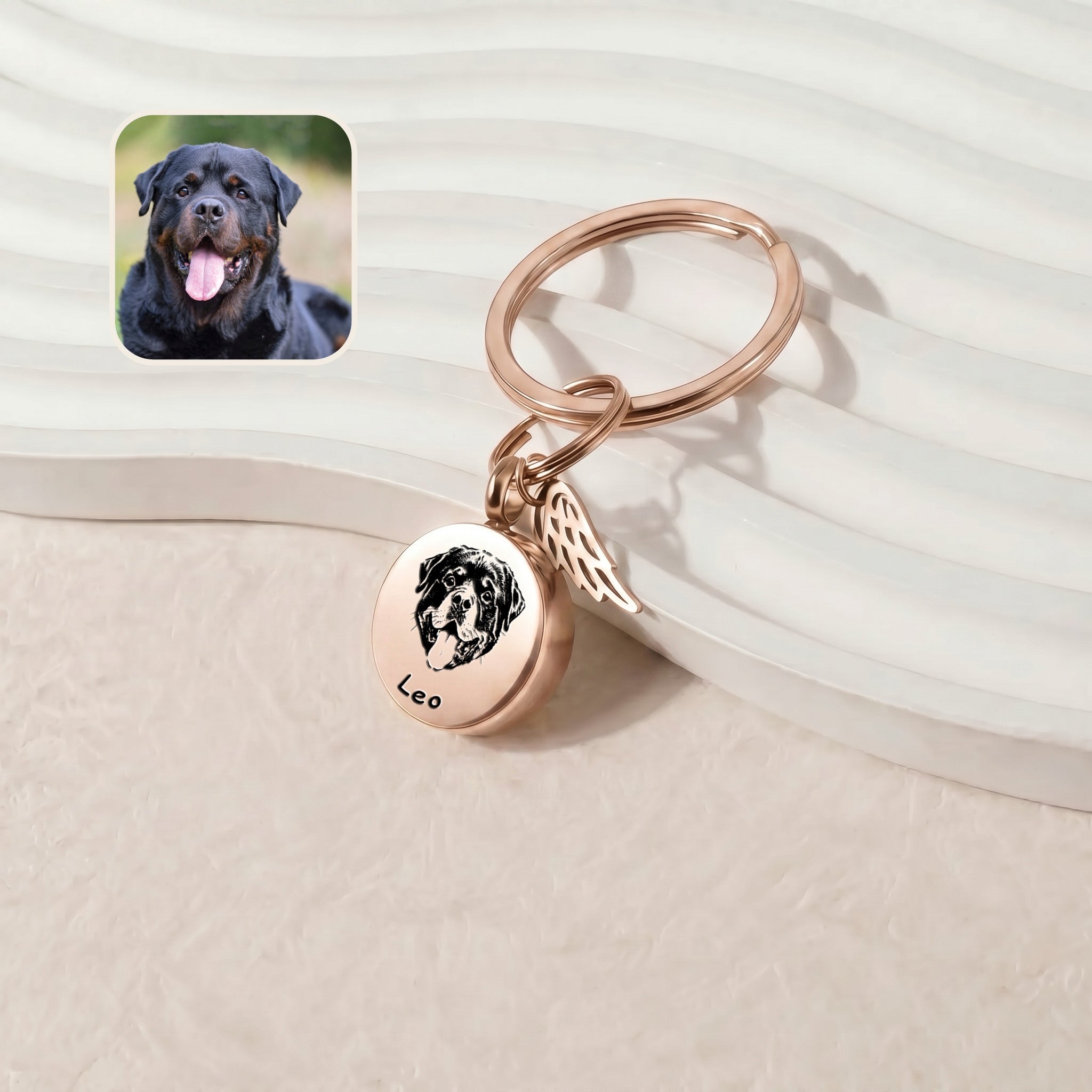 18k rose gold-plated pet cremation disc keychain &mdash; hand-drawn portrait engraved on a 20mm screw-top disc with permanently-attached angel-wing memorial charm, leaned against a ribbed ceramic tray on a cream plush surface beside a reference dog photo inset