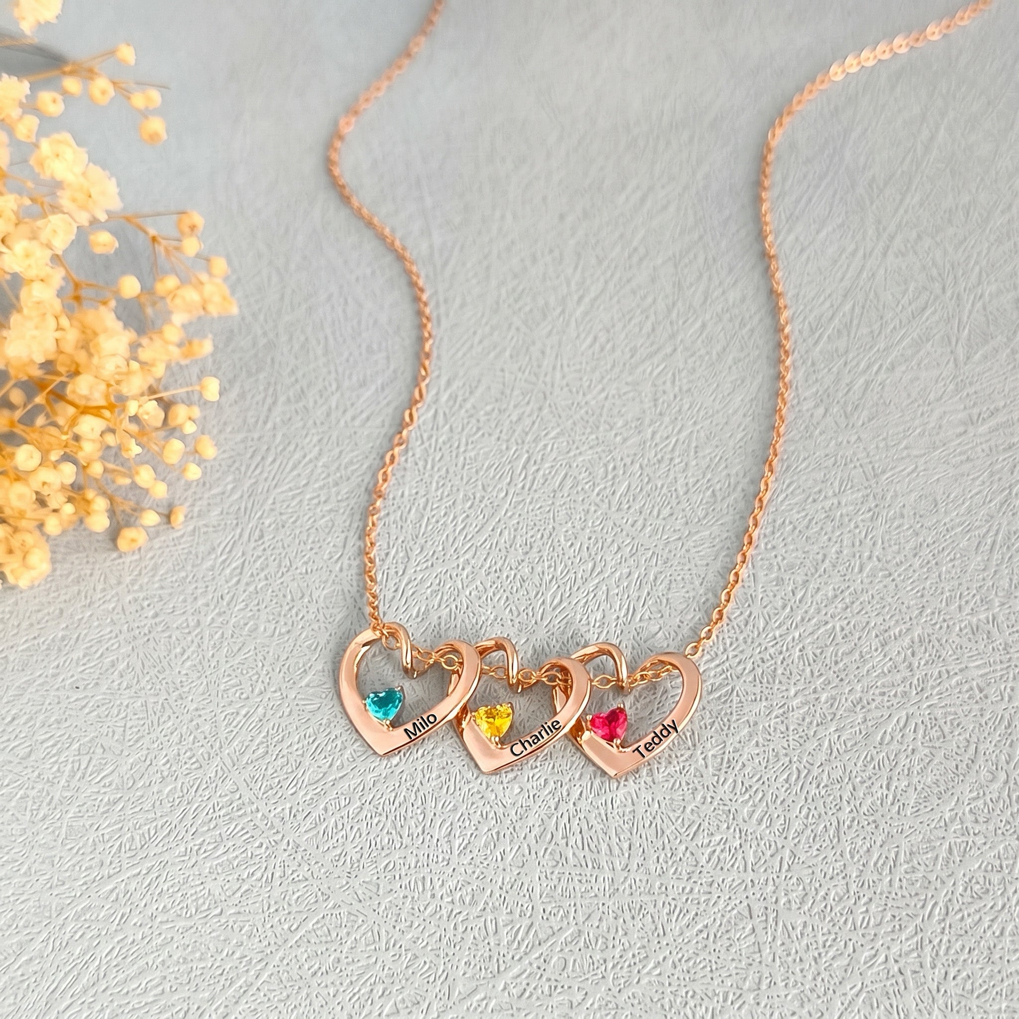 Three rose gold heart pendants with colorful birthstones and pet names on cream background