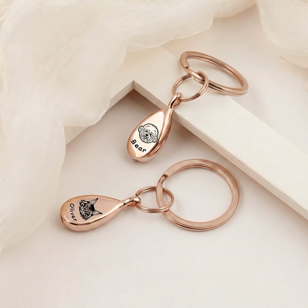Pair of 18k rose gold-plated pet portrait ashes teardrop keychains arranged on a cream plinth under soft gauzy fabric &mdash; both pendants engraved with hand-drawn pet portraits and names