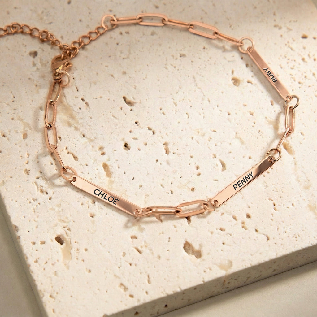 Rose gold paperclip chain pet name bracelet with three engraved name bars on a cream stone block