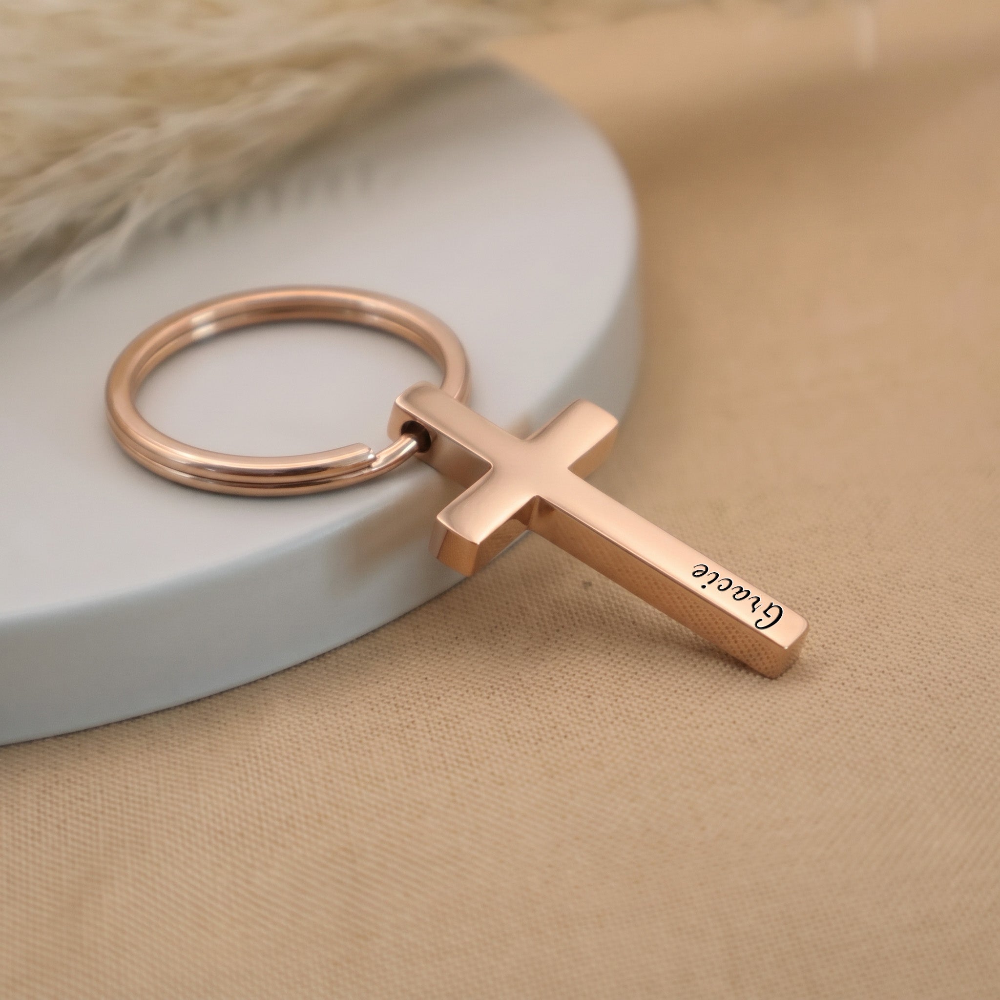 18k rose gold pet cross keychain angled on a ceramic tray with beige fabric — full pendant and 25mm key ring visible in everyday-carry scene