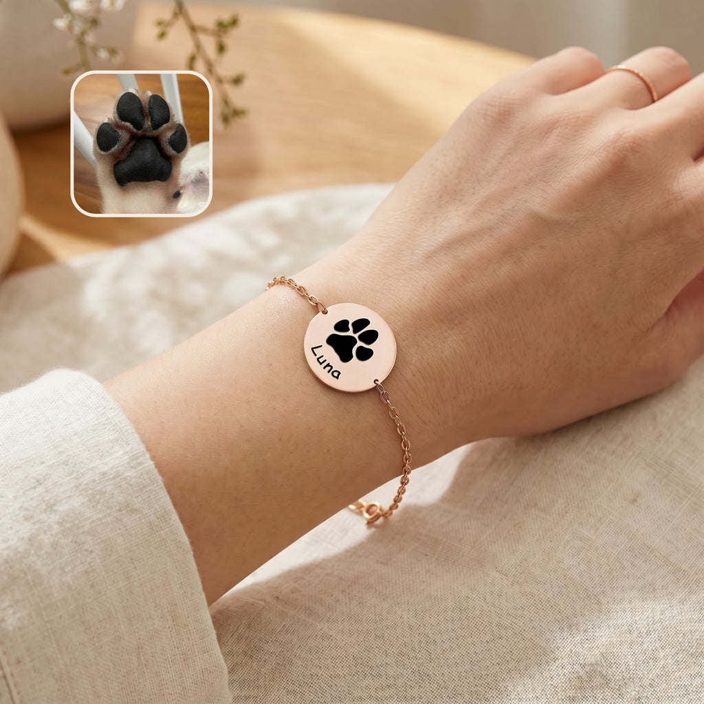 Rose gold chain bracelet on a wrist showing paw print engraving and name on disc charm, linen background