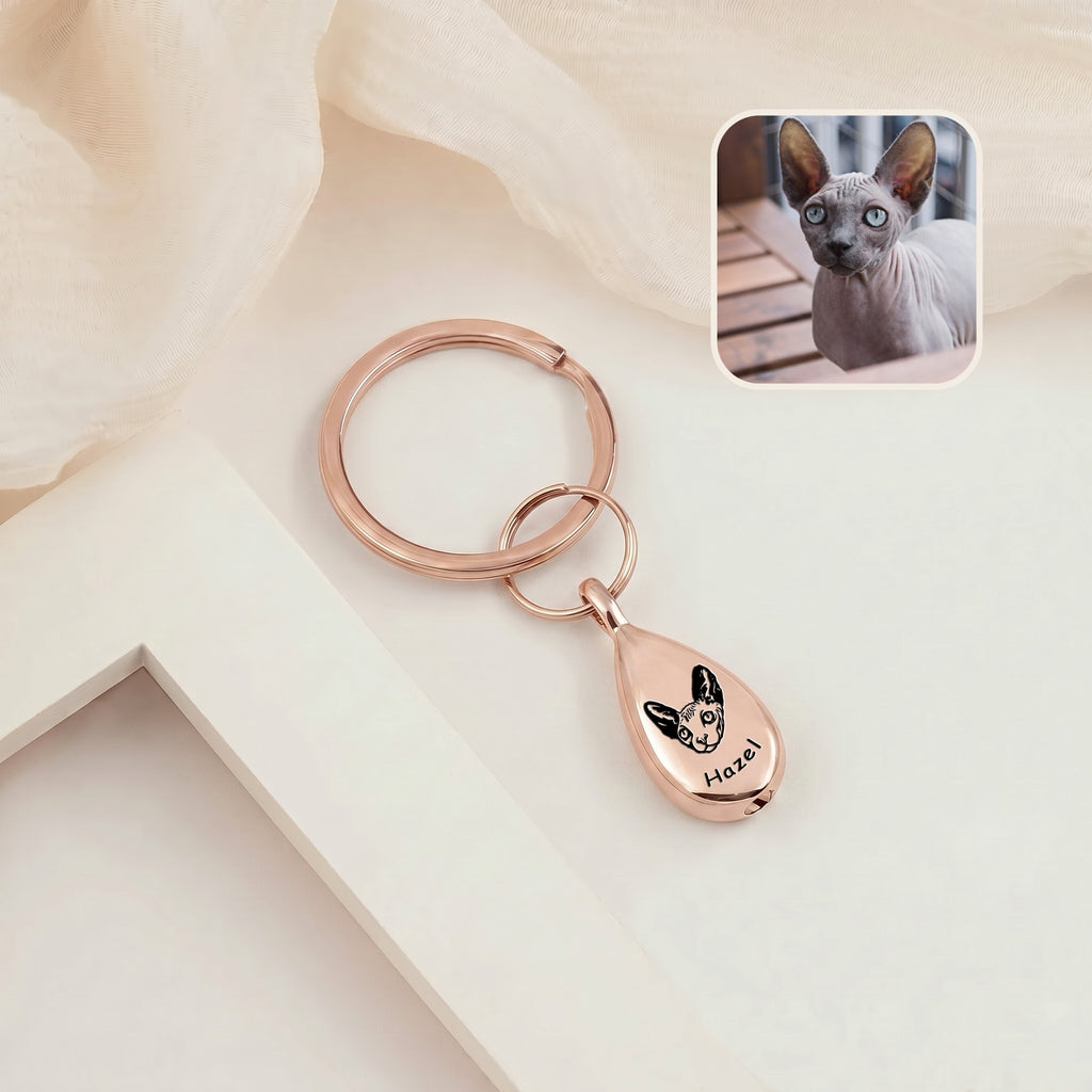 18k rose gold-plated pet portrait ashes teardrop keychain on a cream ledge with sheer fabric &mdash; engraved with a hand-drawn cat portrait and name, with a cat reference photo inset