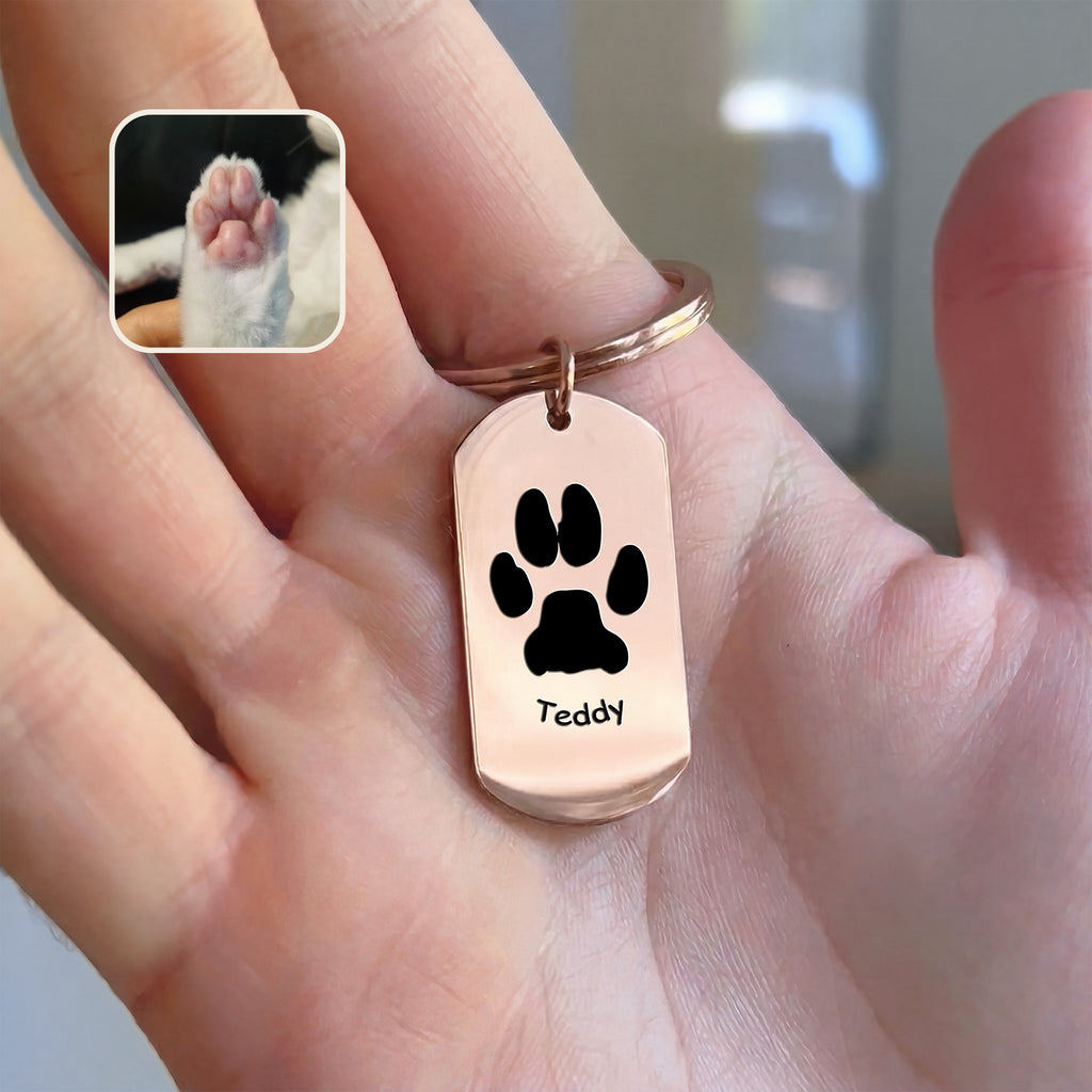 18K rose gold dog tag keychain with paw-print motif and pet name engraving, held in hand beside paw reference photo, warm indoor styled scene