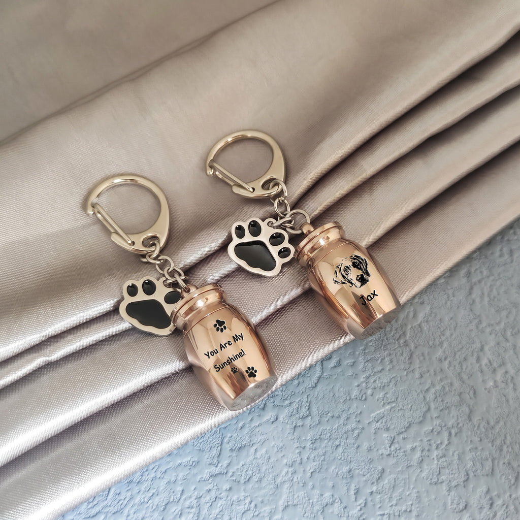 Two 18k rose gold pet urn keychains arranged on draped satin fabric, one engraved with a verse and name, the other engraved with a hand-drawn portrait and name