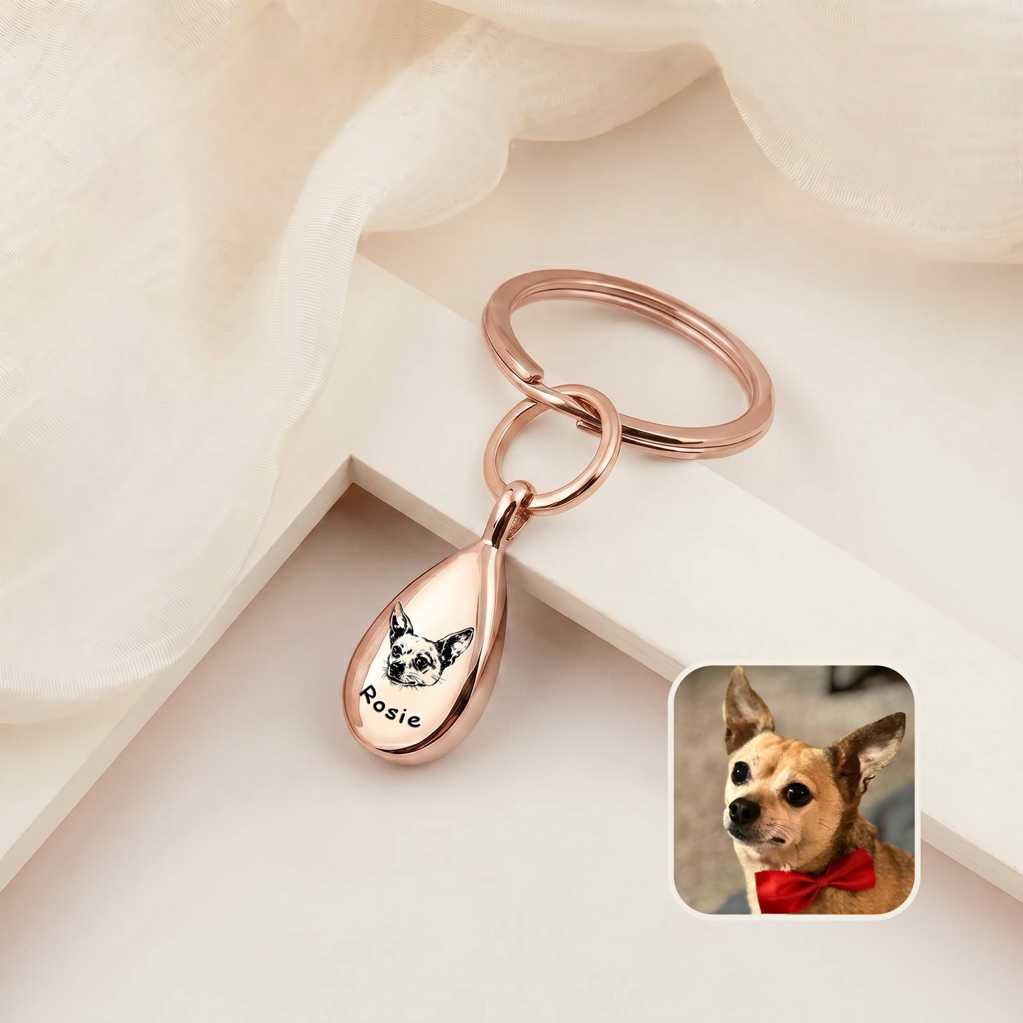 18k rose gold-plated pet portrait ashes teardrop keychain on a cream ledge with sheer fabric &mdash; engraved with a hand-drawn dog portrait and name, with a dog reference photo inset